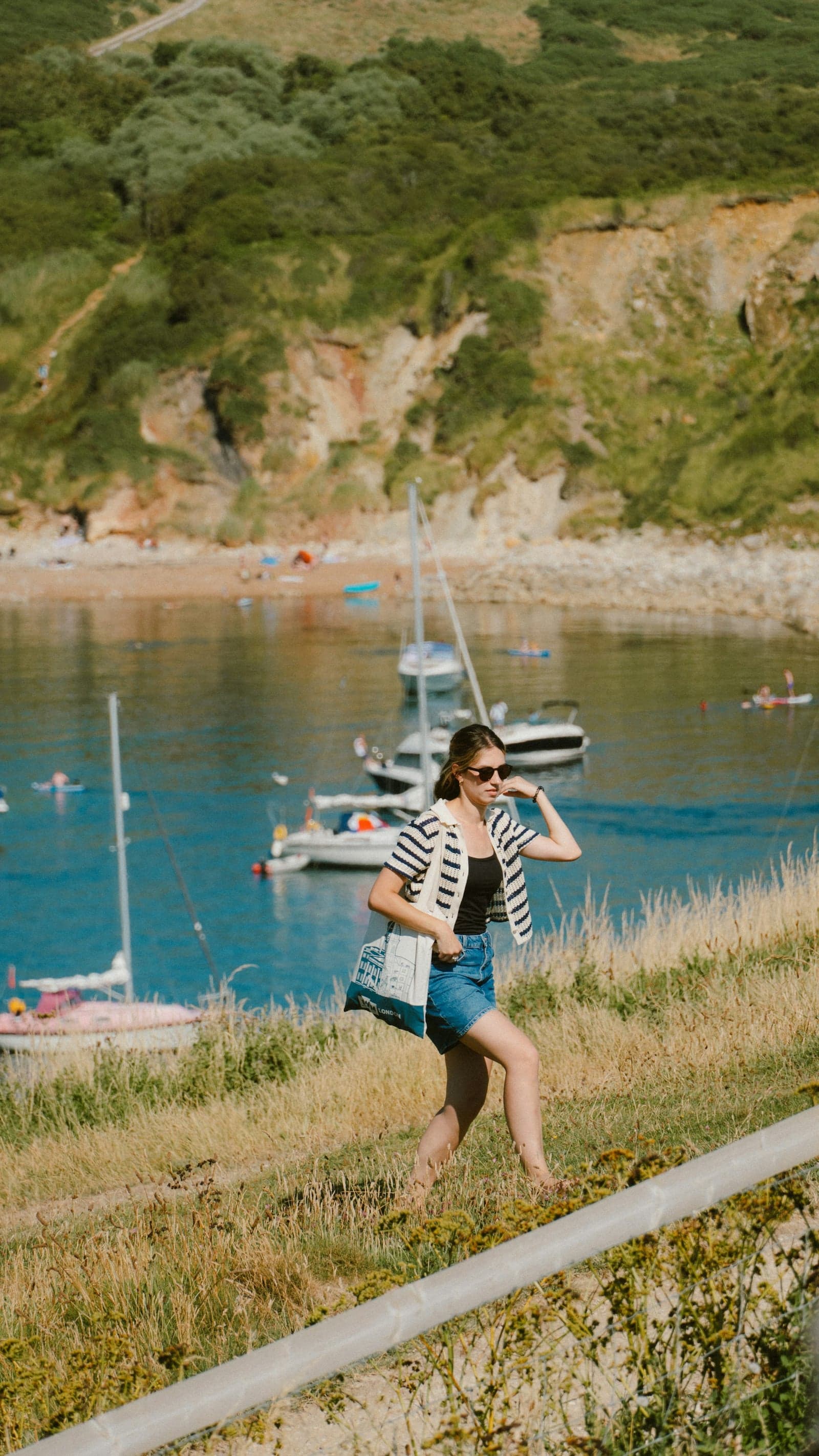 A woman walking a grassy coastal path above a cove of moored sailboats